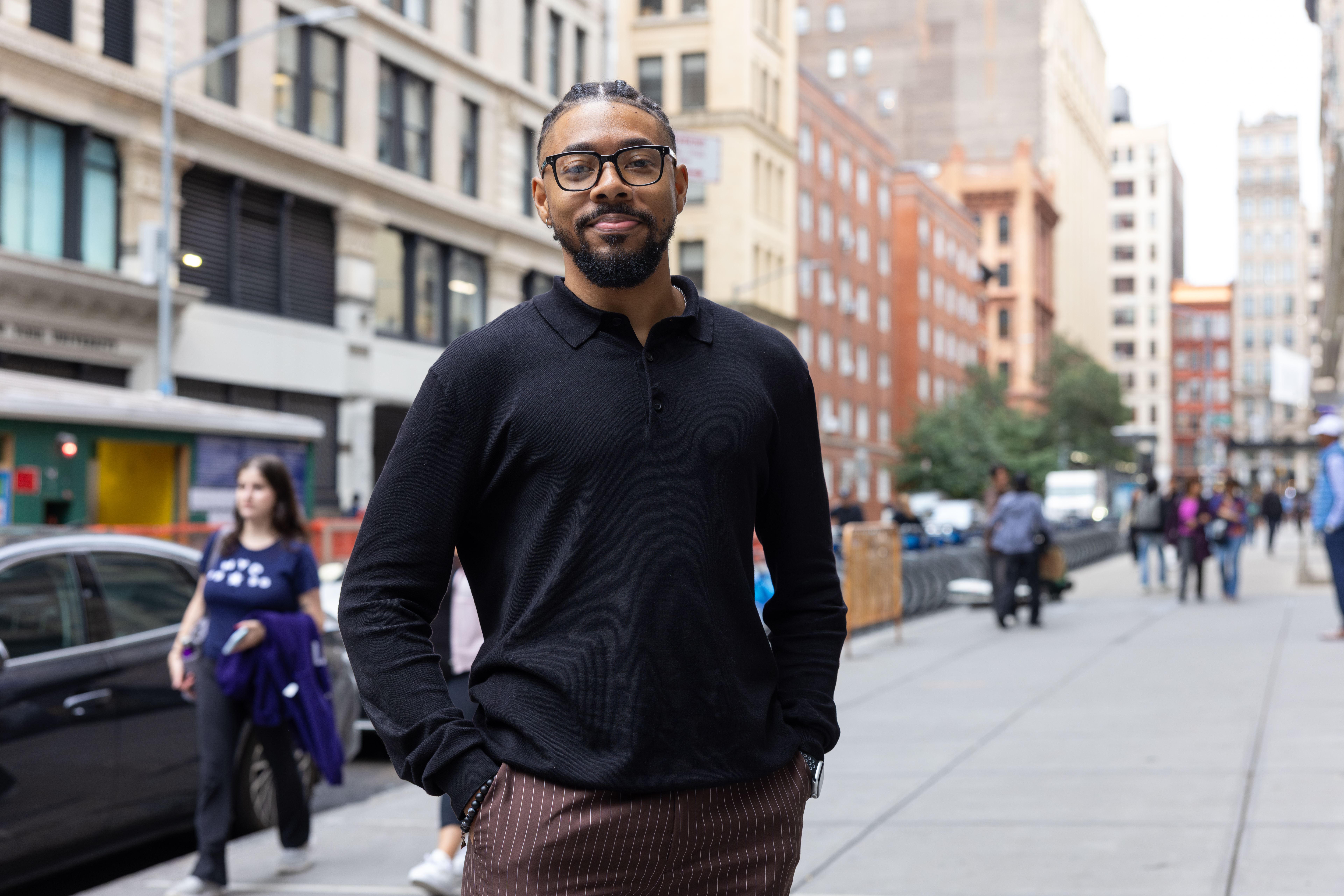 Man stands with hands in his pockets and smile for photo on a city sidewalk. Tall buildings are in the background. People walk in the background.